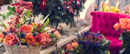 A wicker basket filled with orange roses, green leaves, and red berries sits on a table in a festive, decorated room with a pink tufted sofa and a Christmas tree.