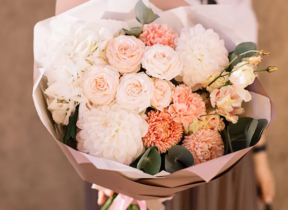 A bouquet of pink, peach, and white flowers, including roses and dahlias, is being held with both hands against a blurred brown background.