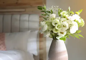 A vase of white and green flowers sits on a bedside table in a softly lit bedroom with a wooden headboard and white bedding.