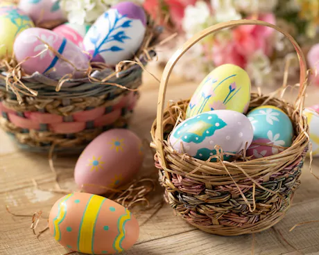 Woven Easter baskets filled with hand-painted pastel eggs rest on a rustic wood table with spring flowers and straw. Each egg is decorated with cheerful floral and spring patterns.