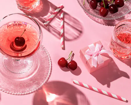 A small gift box, cherries, and charming red cocktails sit on a pink table, showing a beautiful Galentine's day setup