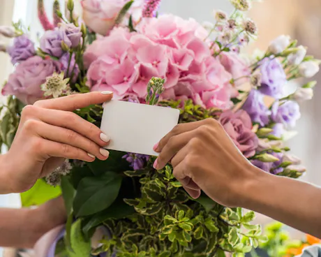 Hands exchanging a blank card in front of a pink and lavender floral arrangement with hydrangea and roses