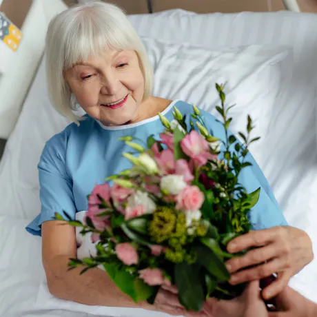 Smiling older woman in a hospital bed receiving a pink and white flower bouquet as a thoughtful get well gift