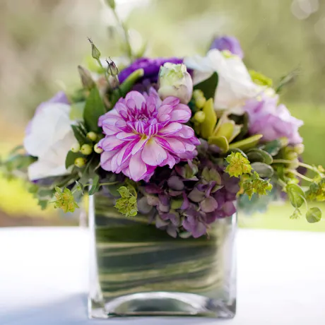 Purple, green and white floral arrangement with hydrangea, dahlia, lisianthus, greenery, and small yellow accent flowers in a glass vase