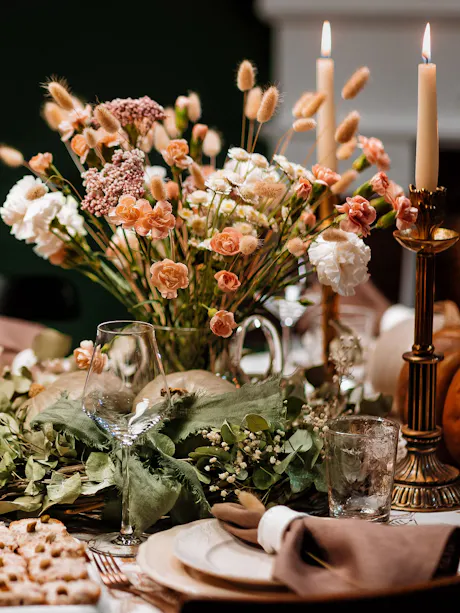 Autumn dinner table centerpiece with peach and white flowers, dried grasses, greenery, glowing taper candles, glassware, and neutral place settings