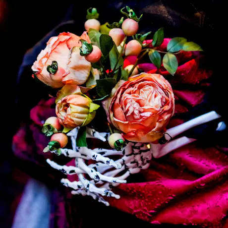 Halloween flower arrangement with peach roses, decorative berries, dark red flowers, greenery, clasped in prop skeleton hands
