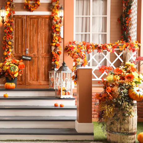 Fall front porch decorated with orange floral arrangements, pumpkins, autumn leaf garlands, lanterns, and seasonal Halloween d&eacute;cor