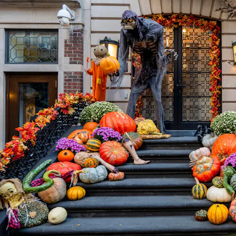 Halloween front steps decorated with pumpkins, gourds, purple and white mums, a giant skeleton figure, and spooky seasonal props
