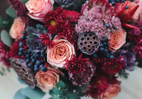 A bouquet of flowers displaying pink roses, crimson chrysanthemums, purple asters, berry clusters, and lotus seed pods, arranged together against a blurred green foliage background.