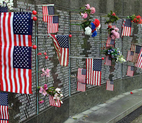 American flags and bouquets of flowers adorn a black granite memorial wall engraved with numerous names, honoring fallen soldiers. This solemn tribute creates a poignant atmosphere of remembrance and respect.