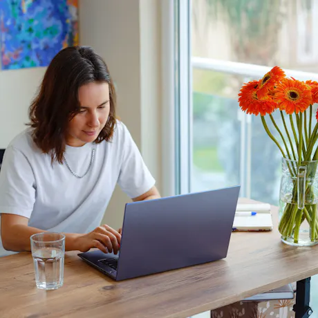 Person working on a laptop beside a vase of orange gerbera daisies in a bright home office