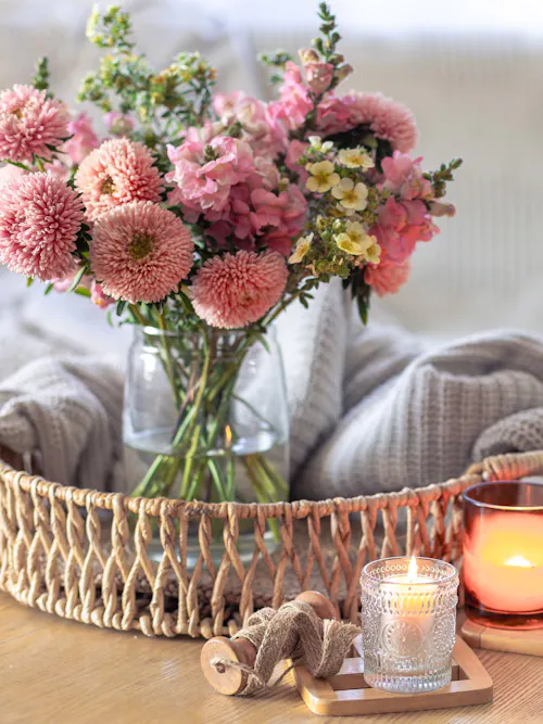 Pink chrysanthemum arrangement in a clear glass vase beside glowing candles and blankets on a wooden table.
