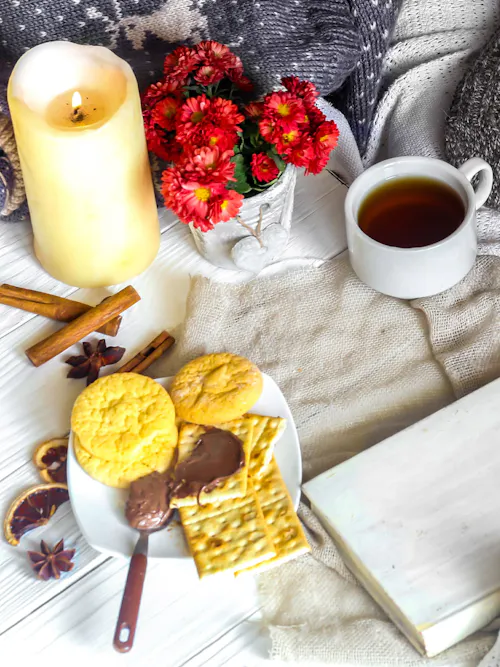 Red chrysanthemums beside a glowing candle, cup of tea, cookies, cinnamon sticks, blanket, and white book making up a winter setting.