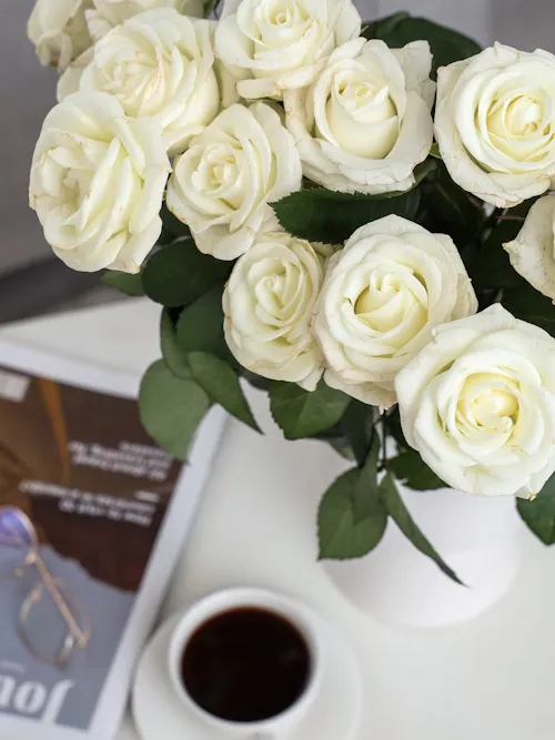 Close-up of fresh white roses in a white vase beside a cup of coffee, creating a simple home floral display.